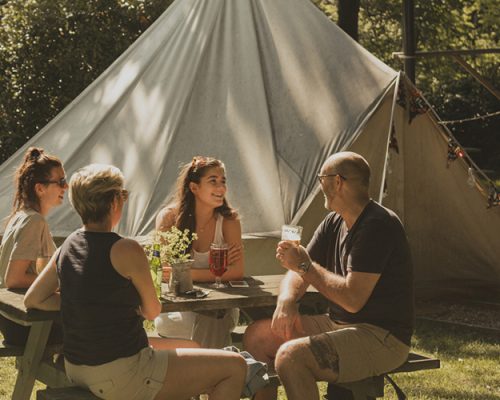 Family of four are sitting at a picnic bench next to their glamping tent. They are all talking and enjoying an alcoholic beverage.