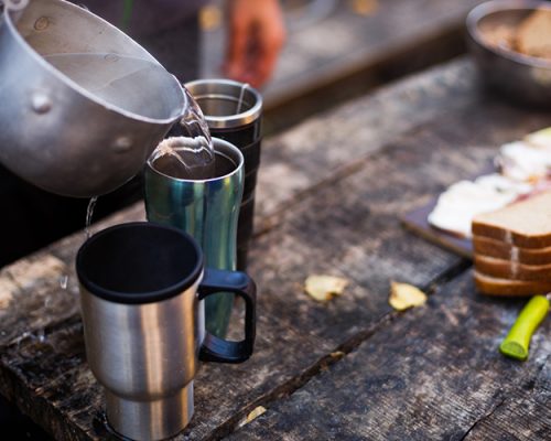 Man pour boiled water to hot mugs on wooden table during breakfast at the forest camp. People camping at national park and doing lunch tea and coffee.