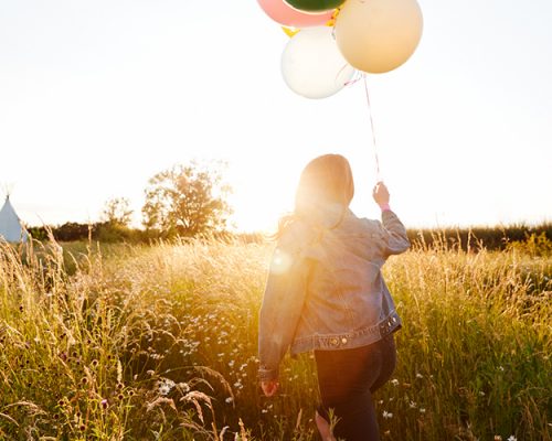 Rear View Of Woman Camping At Music Festival Running Through Field With Balloons Against Flaring Sun